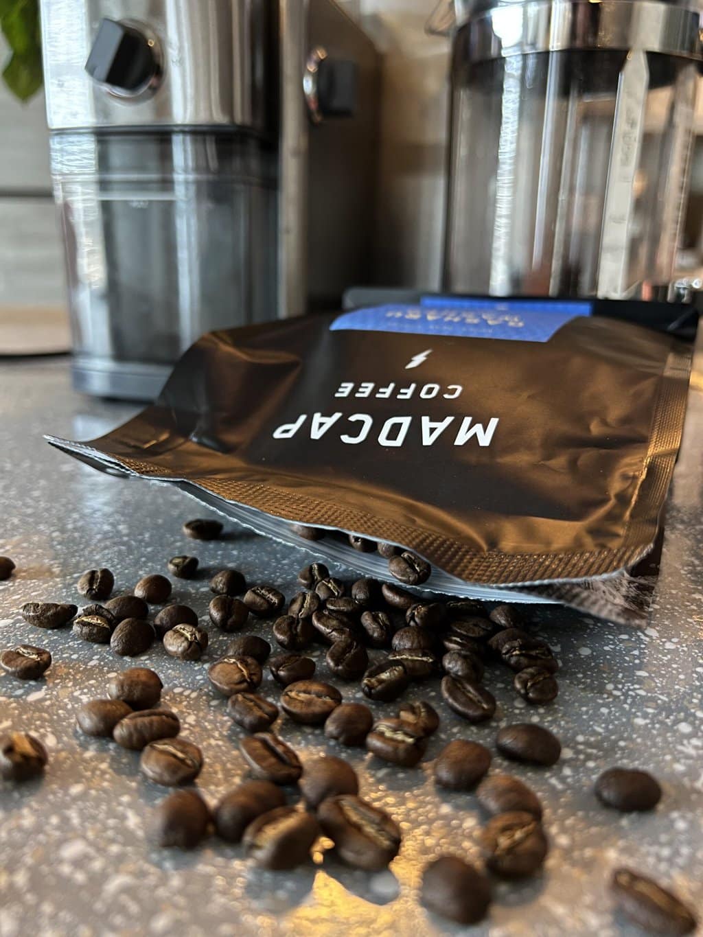 Madcap coffee pack and scattered coffee beans against the background of a coffee grinder and a french press