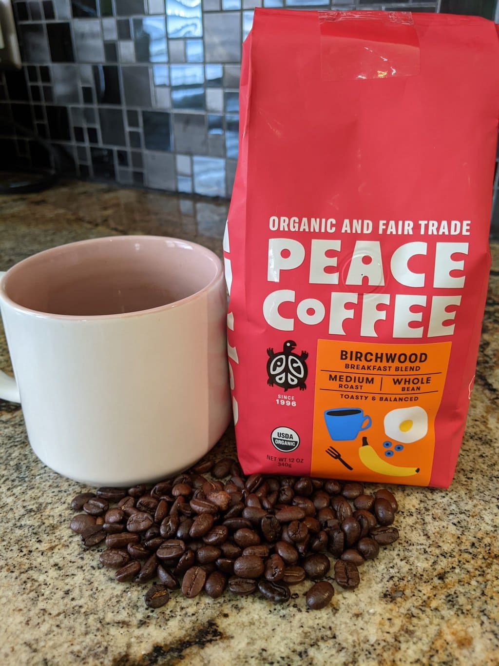 Peace Coffee Birchwood Breakfast Blend Medium Roast stands on the kitchen table next to a brewed cup of coffee and scattered coffee beans