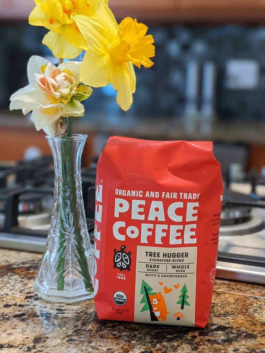 On the kitchen table, next to a vase with daffodils, there is a package of Peace Coffee Tree Hugger Signature Blend Dark Roast